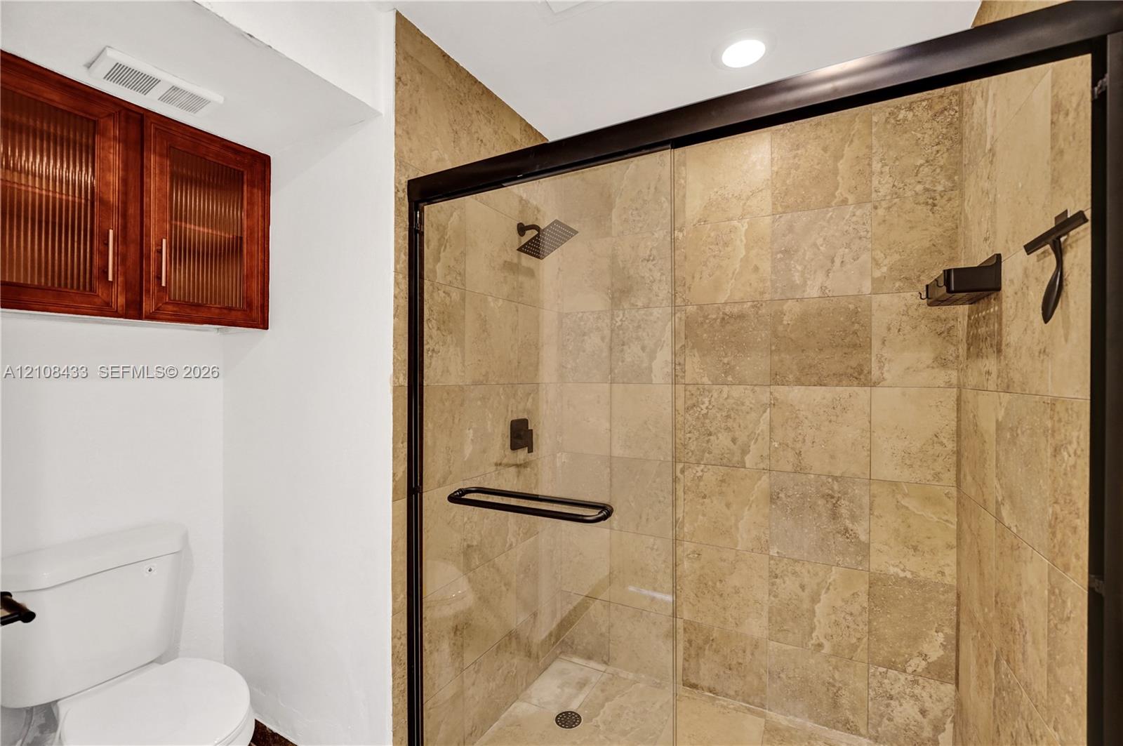 Walk-in shower with natural stone tiling, matte black frame, glass door, and overhead rainfall showerhead, complemented by wall-mounted wood-finish cabinetry