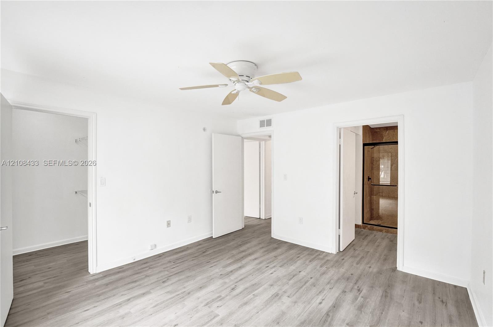 Neutral interior featuring wood-finish flooring, a ceiling fan with integrated lighting, and multiple interior doorways leading to a walk-in closet and a bathroom with a glass-enclosed shower