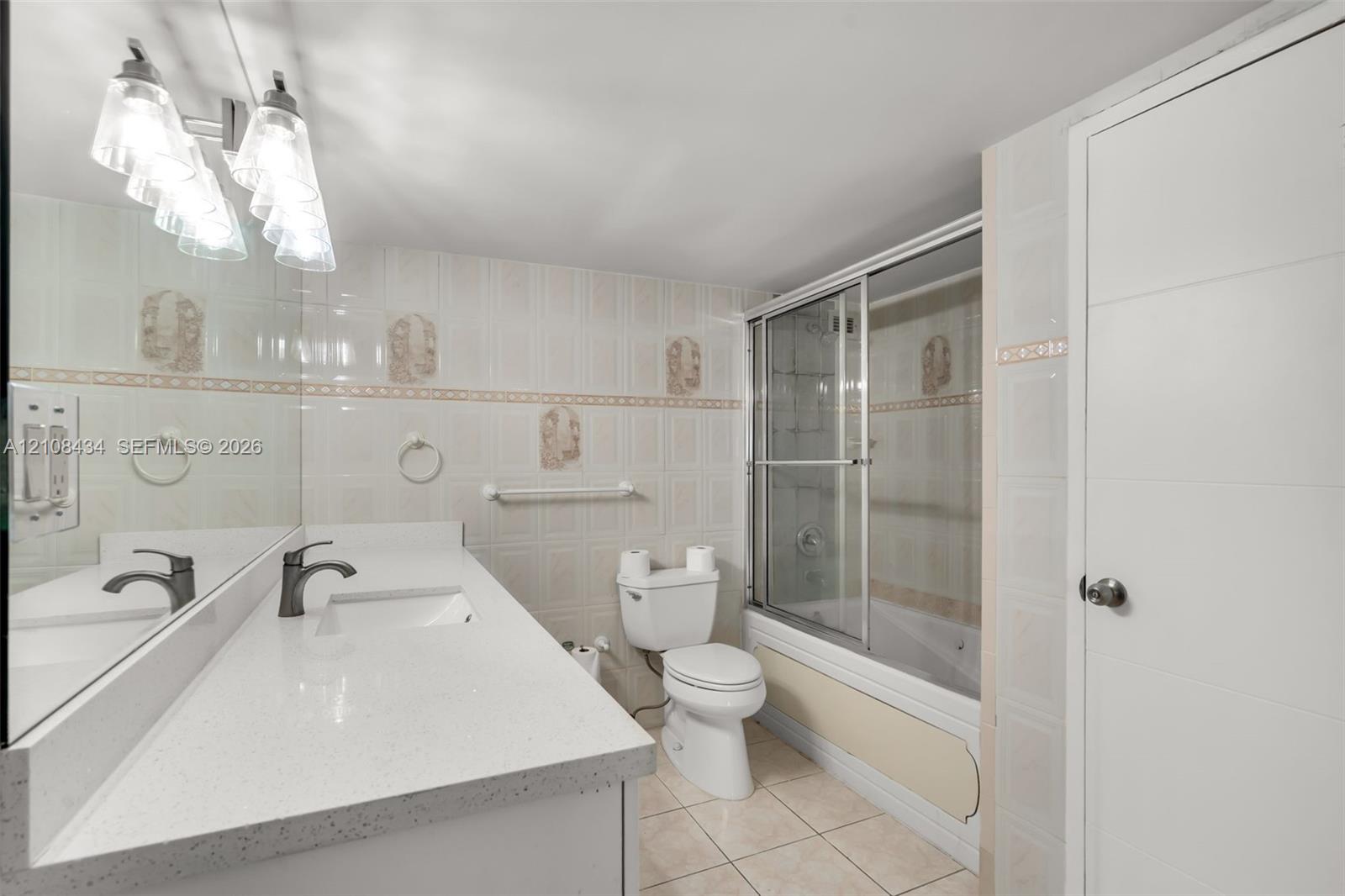 Bathroom featuring a quartz-topped vanity with an integrated basin, ceramic tile flooring, full-width wall mirror, and a tub-shower combination enclosed by sliding glass doors