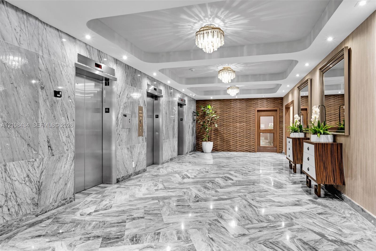 Polished stone flooring in a herringbone pattern, full-height stone wall cladding, stainless steel elevator doors, recessed tray ceiling with multi-tiered crystal chandeliers, and textured wood-paneled accent walls