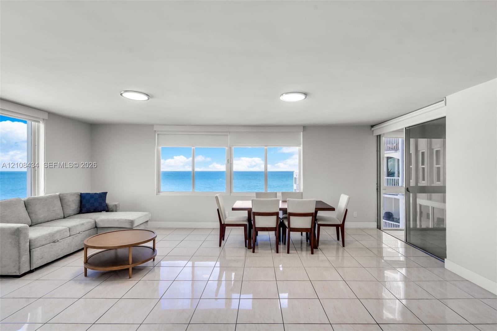 Open-concept room featuring light-toned tile flooring, large multi-pane windows, and a sliding glass door providing access to a balcony with an ocean backdrop