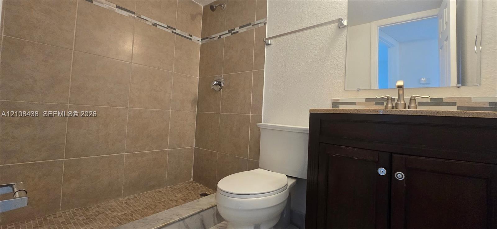 Bathroom featuring a vanity with a countertop, tiled walk-in shower with decorative inlay, wall-mounted mirror, and metallic finish hardware