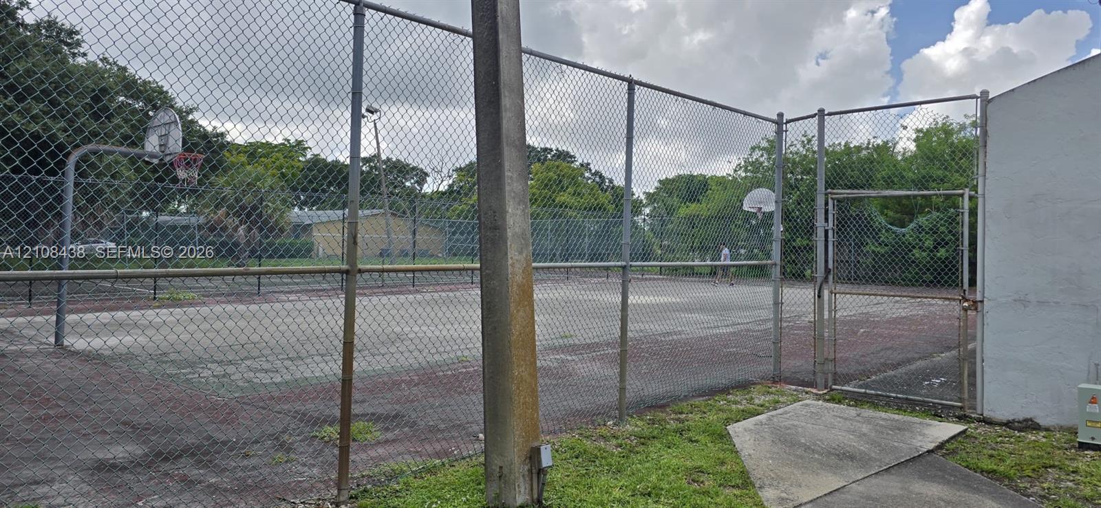 Recreational court featuring a hard surface, perimeter chain-link fencing, dual basketball hoops, and a gated entrance