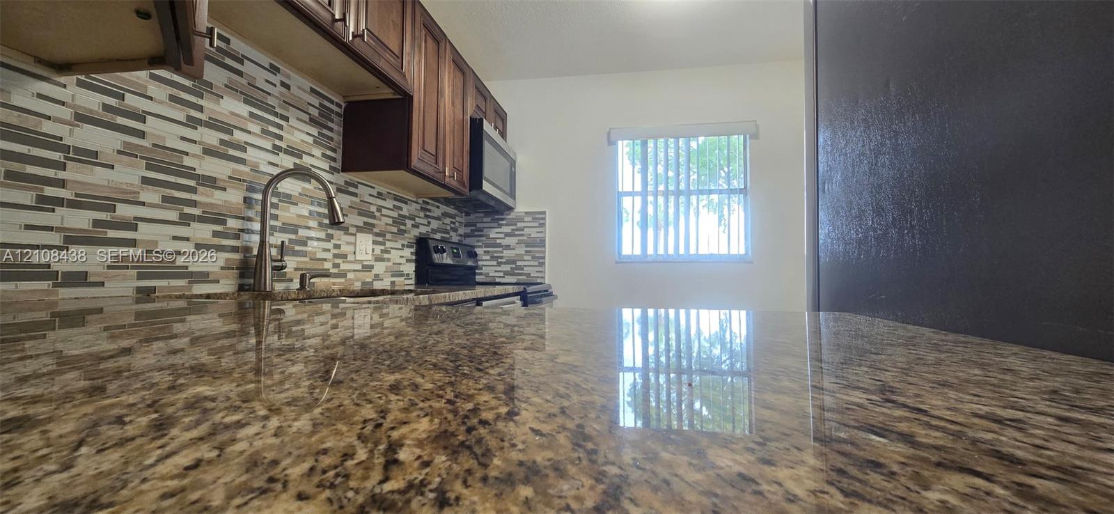 Kitchen featuring stone-finish countertops, multi-toned linear tile backsplash, dark wood-finish cabinetry, built-in microwave, and arched faucet