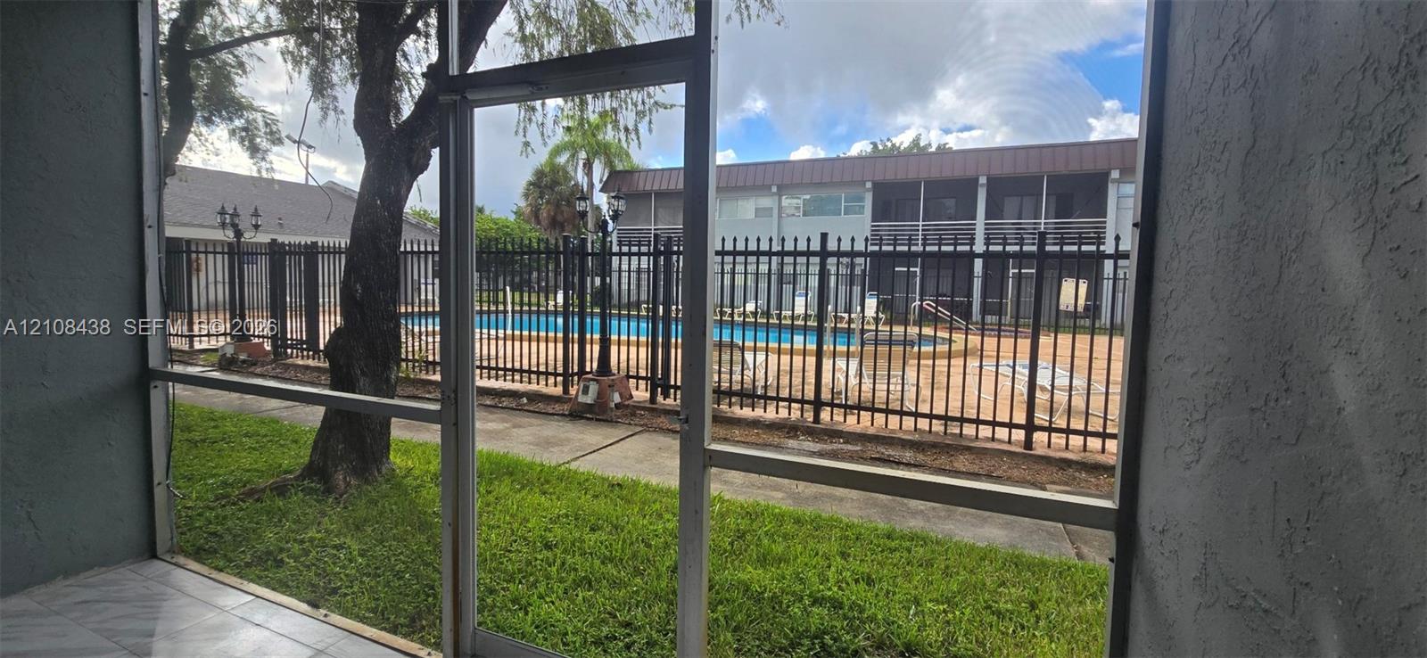 Screened-in patio with tile flooring, featuring proximity to a community swimming pool enclosed by black metal fencing and mature landscaping