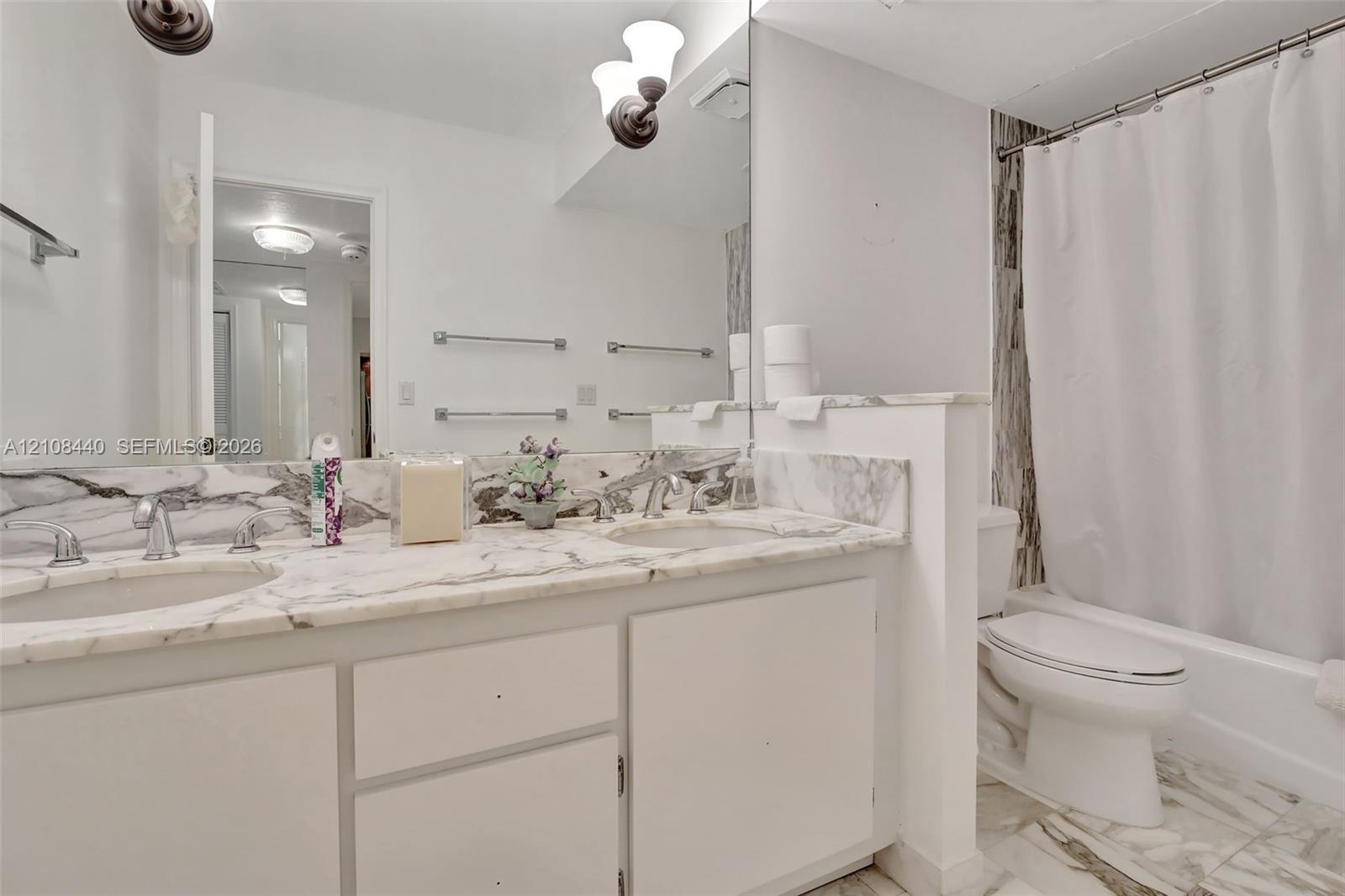 Dual-sink vanity with stone-finish countertop and backsplash, wall-to-wall mirror, brushed metal hardware, and tub-shower combination with floor-to-ceiling stone-finish surround