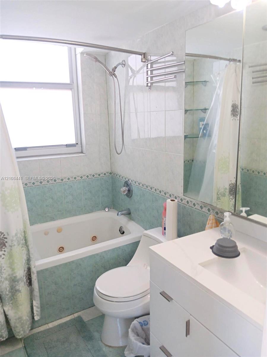 Bathroom featuring a jetted tub, wall-mounted handheld shower fixture, modern white vanity with a rectangular integrated sink, large vanity mirror, and glass wall shelving