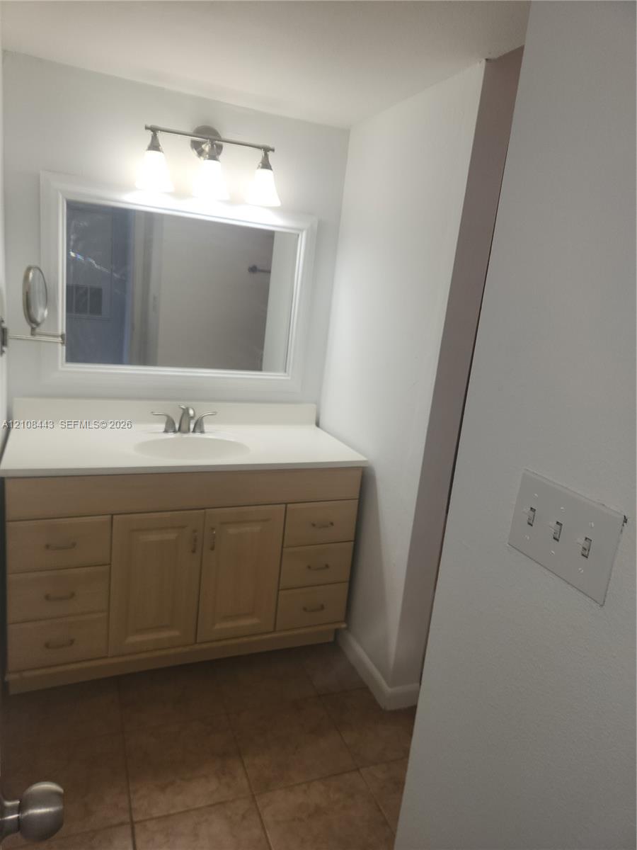 Light-toned wood vanity with ample drawer storage, integrated sink, large wall-mounted mirror, and three-light fixture over tile flooring