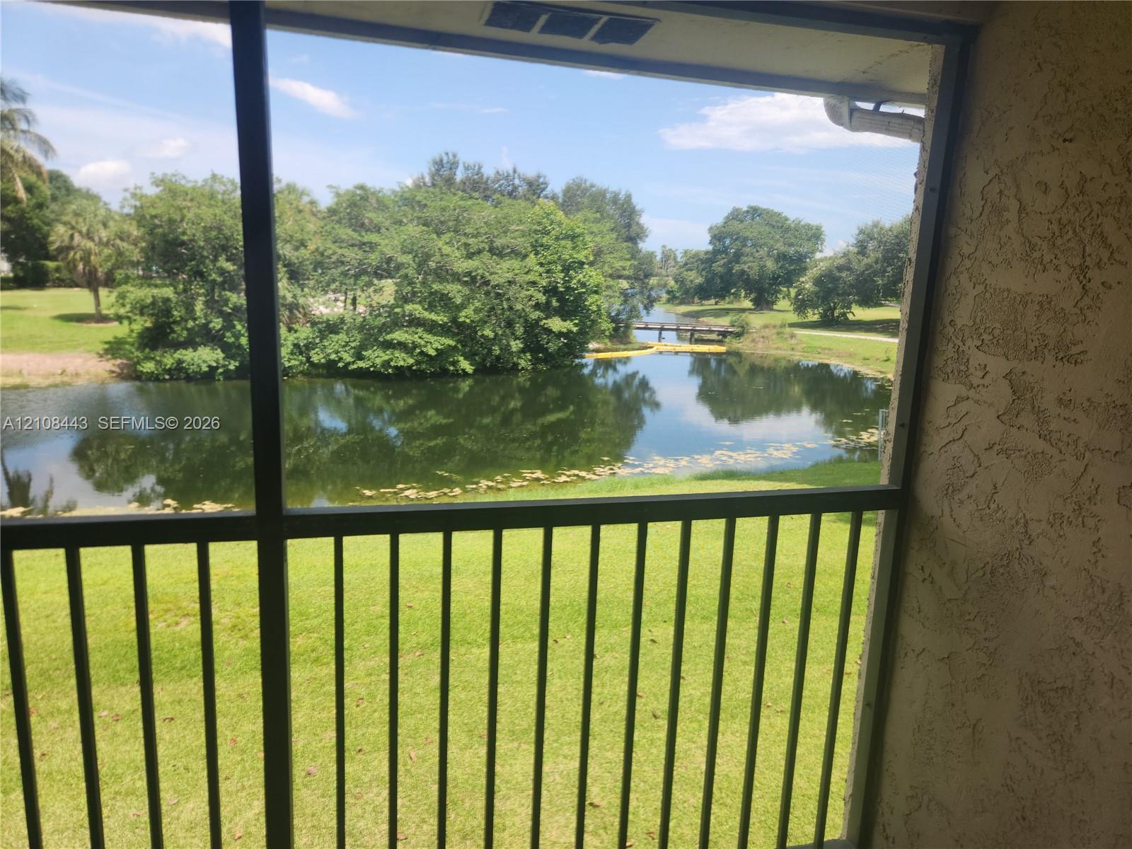 Screened-in balcony with metal railing, overlooking a pond, mature trees, and pedestrian bridge Screened-in balcony with metal railing, overlooking a pond, mature trees, and pedestrian bridge