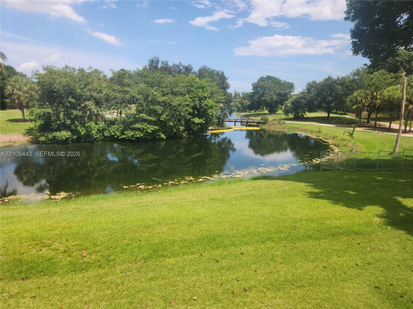 Waterfront pond featuring a grass lawn, mature trees, and metal perimeter fencing