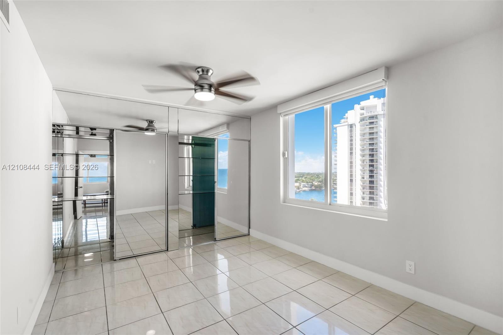 Large format tile flooring, floor-to-ceiling mirrored closet doors, ceiling fan with integrated lighting, and expansive window with water and skyline exposure