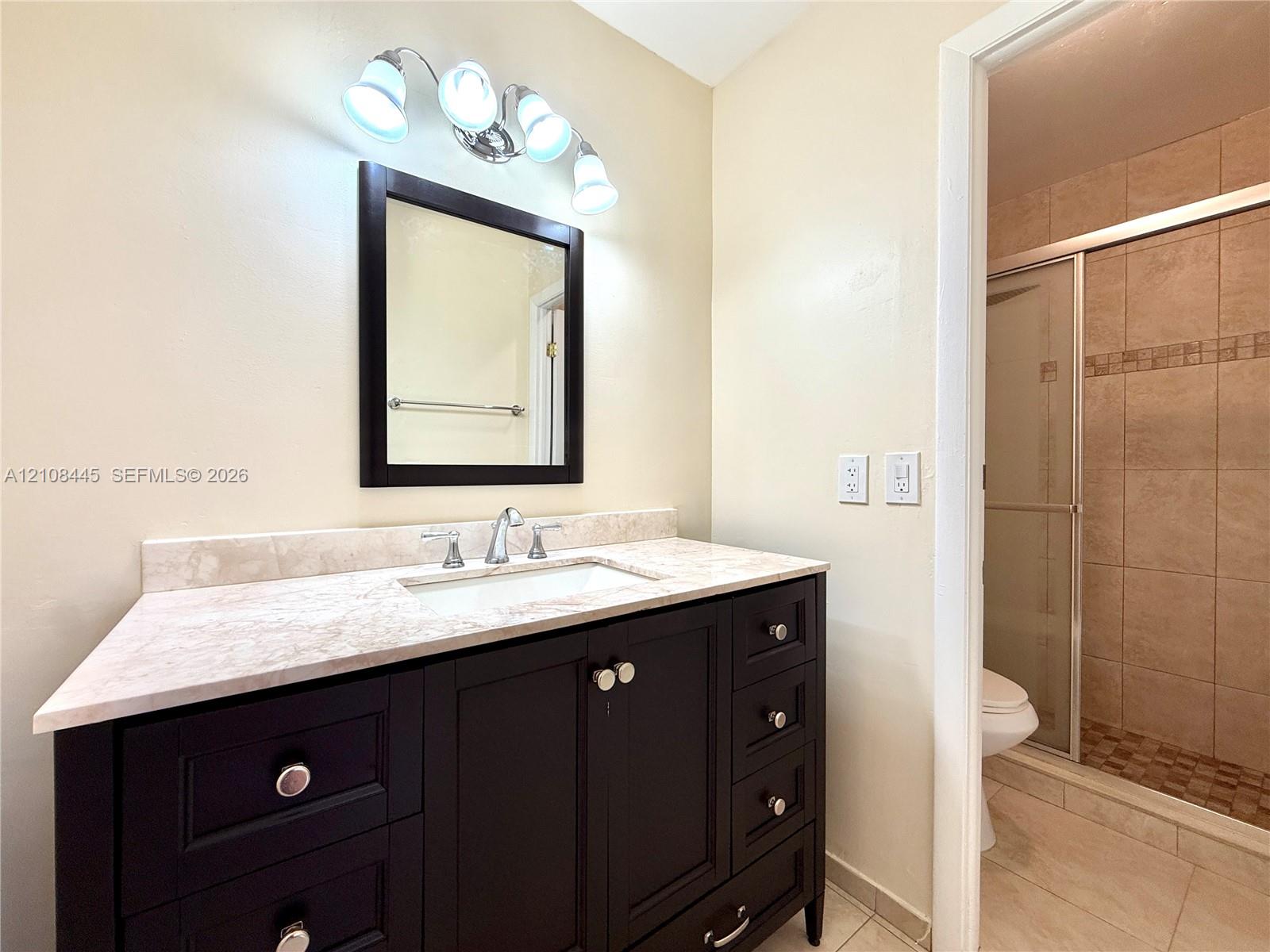 Dark-finish wooden vanity with a stone-top counter, undermount sink, multi-bulb wall lighting, and shower with tiled wall surround