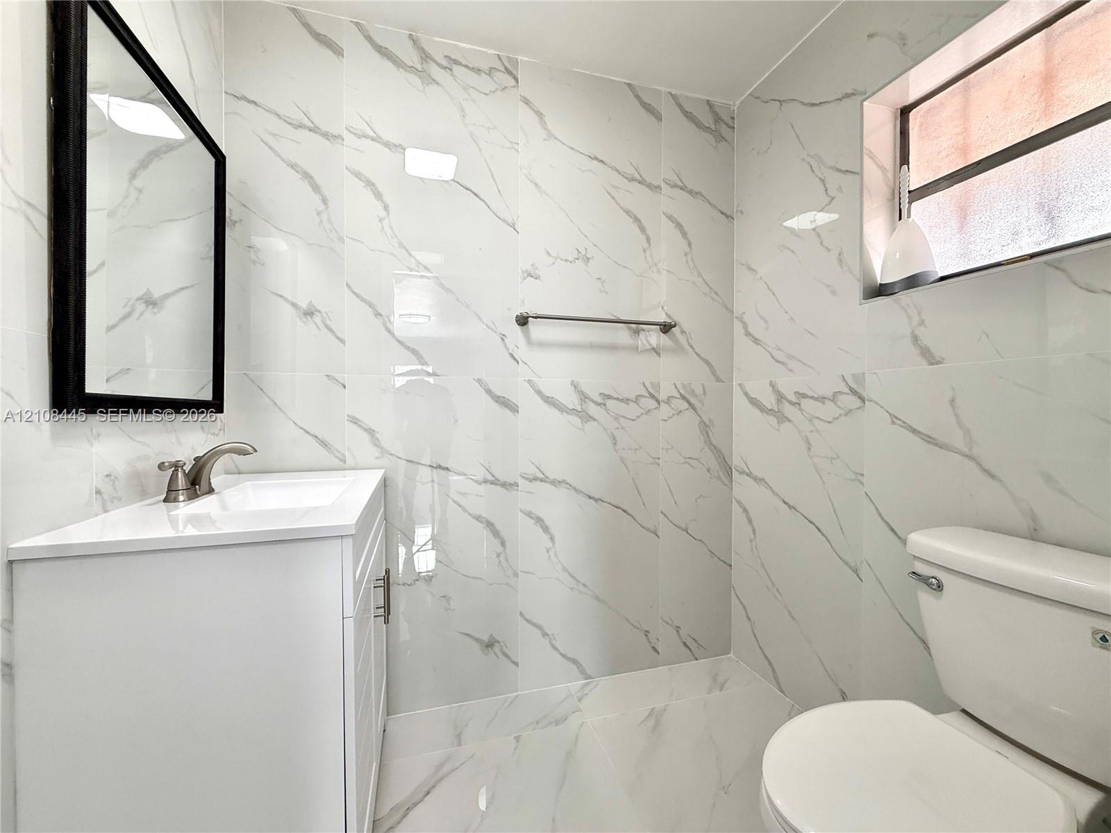 Guest Bathroom featuring white veined wall and floor tile, white vanity cabinet with integrated sink, rectangular black-framed wall mirror, and brushed-finish metal faucet and hardware