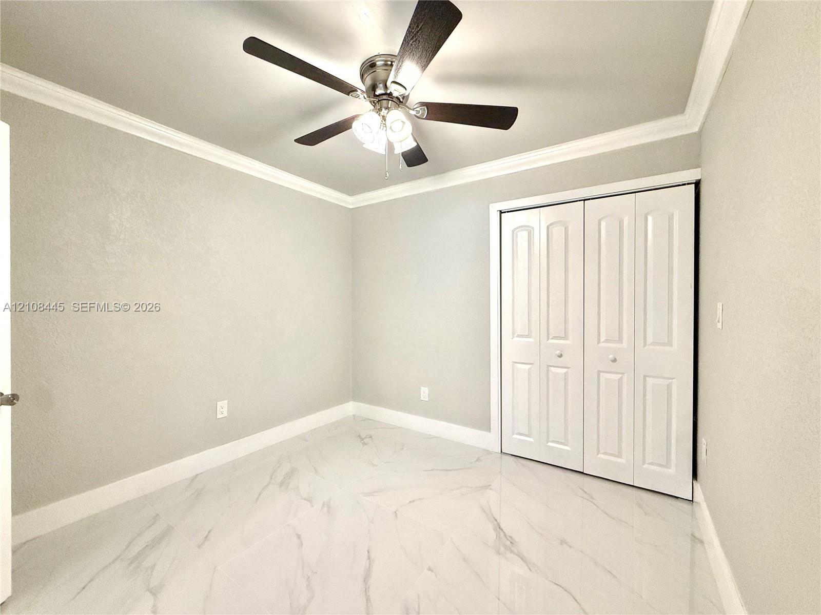 Downstairs Bedroom. Large-format tile flooring with a neutral veined pattern, crown molding, ceiling fan with integrated lighting, and white bi-fold closet doors