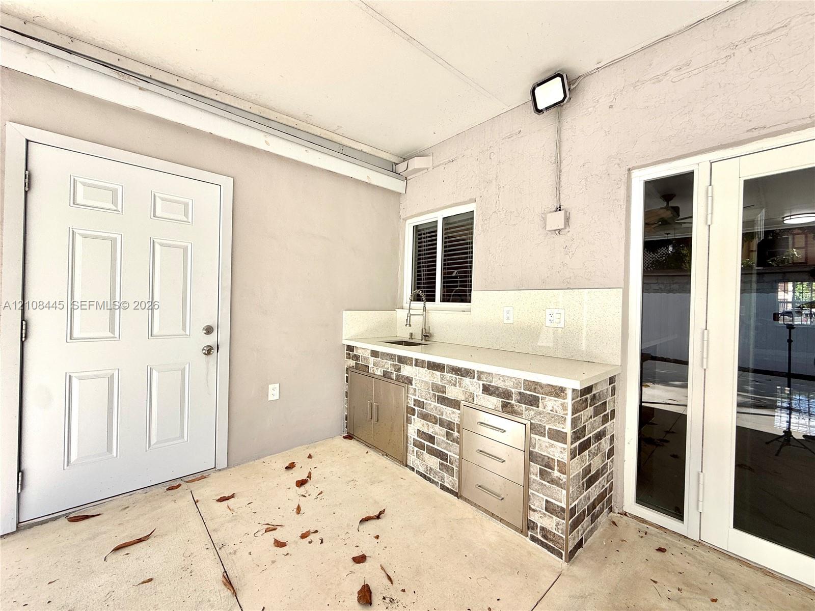 Covered outdoor kitchen area featuring a stone-veneer base, solid surface countertop, stainless steel integrated sink, and built-in metal cabinetry