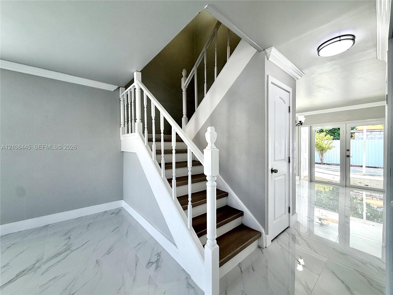 Staircase featuring dark wood-finish treads, white balusters, and a decorative newel post, complemented by polished stone-finish flooring and double-panel glass doors