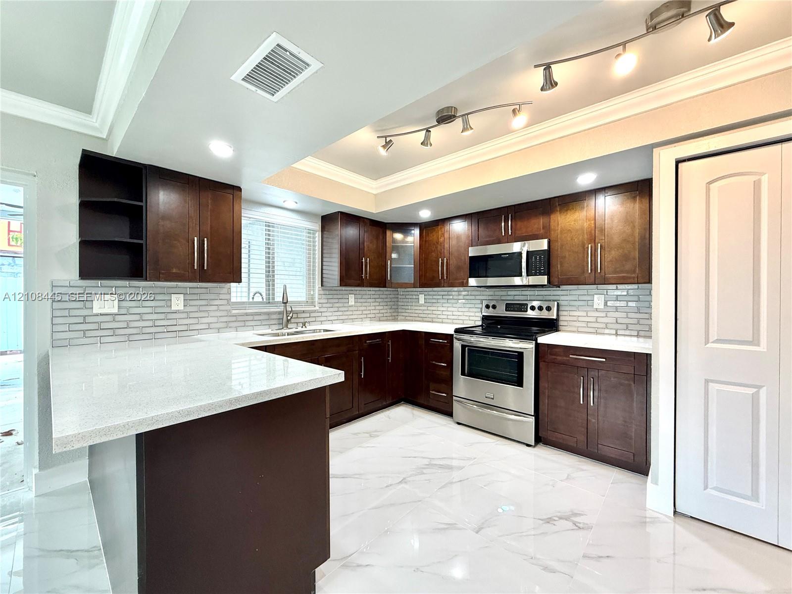 Kitchen featuring dark wood cabinetry, solid-surface countertops, brick-style tile backsplash, stainless steel appliances, and large-format tile flooring with recessed and track lighting