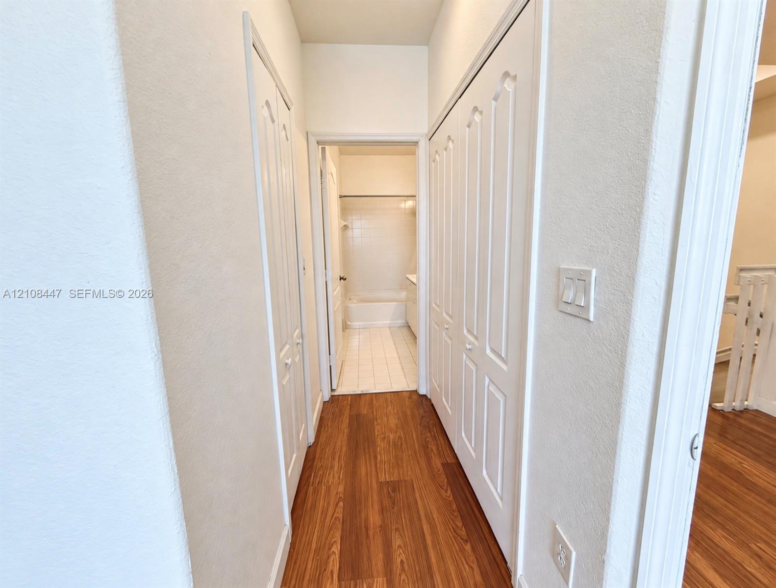 Hallway featuring wood-finish flooring, dual sets of white paneled bi-fold closet doors, and a bathroom entry with white tile flooring and a tub-shower combination