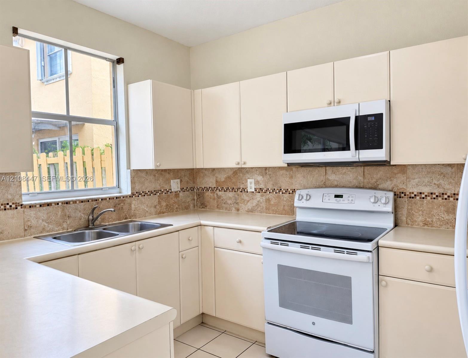 Kitchen featuring light-finish cabinetry, stone-texture tile backsplash with decorative inlay, solid-surface countertops, double-basin sink, and built-in microwave