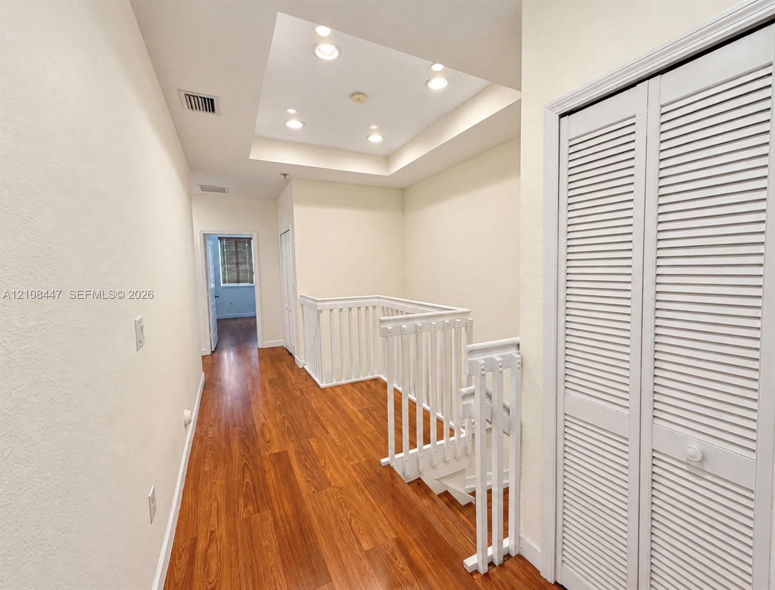 Wood-finish flooring, recessed tray ceiling lighting, white baluster staircase railing, and louvered bifold closet doors
