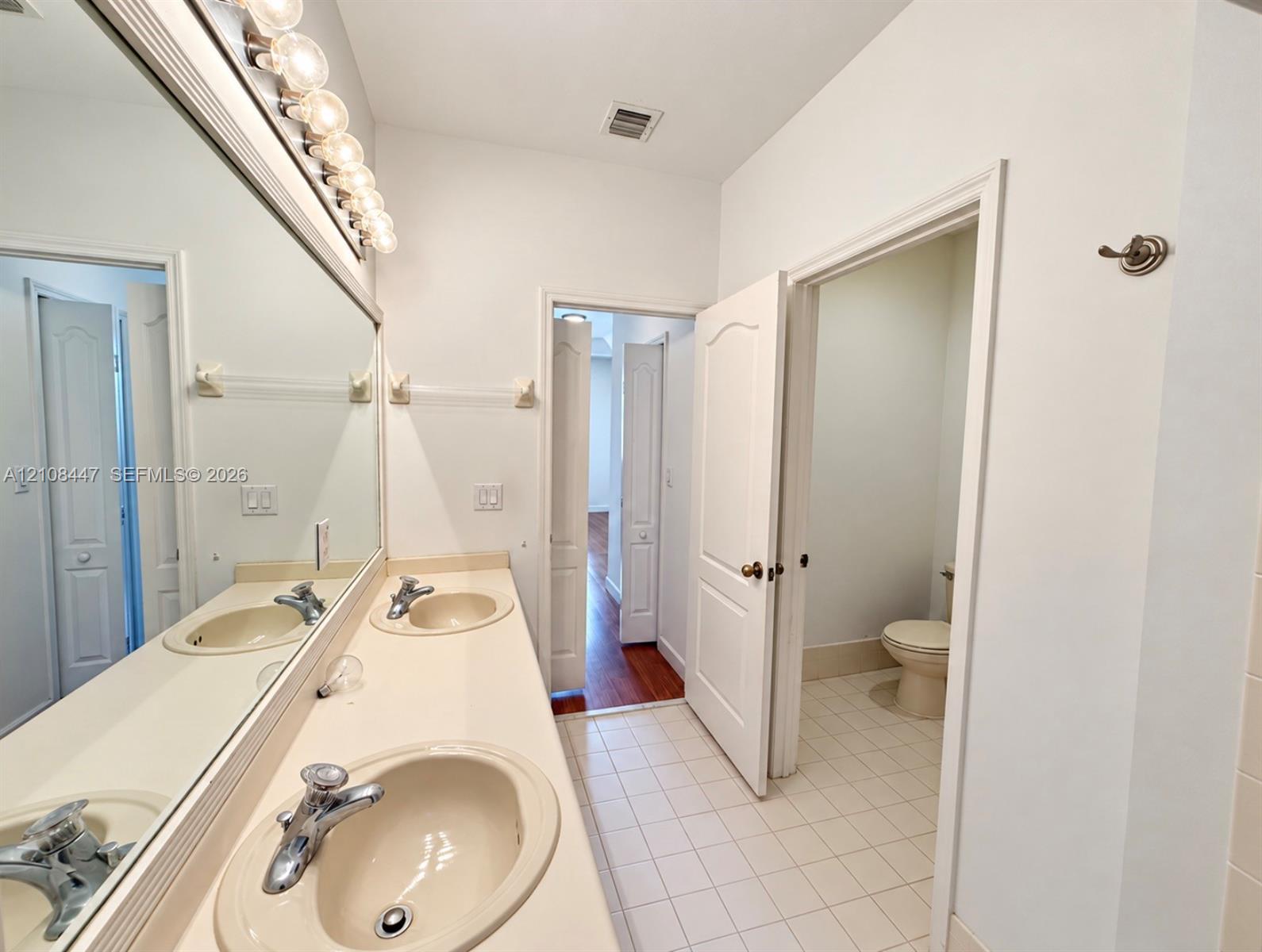 Full-width vanity featuring dual integrated sinks, solid-surface countertop, and expansive wall-mounted mirror with multi-bulb light fixture