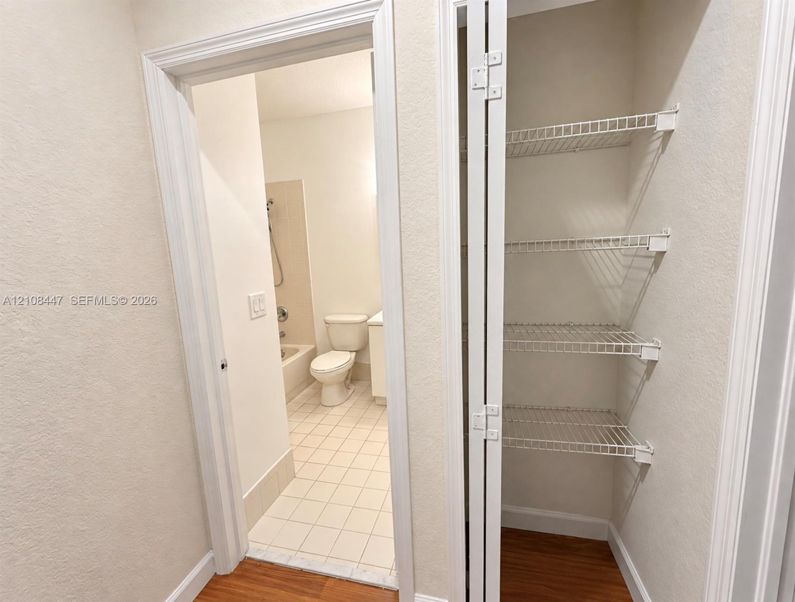 Storage closet featuring white wire shelving and bifold doors, with interior access to a space containing tile flooring, a bathtub-shower combination, a porcelain toilet, and a vanity