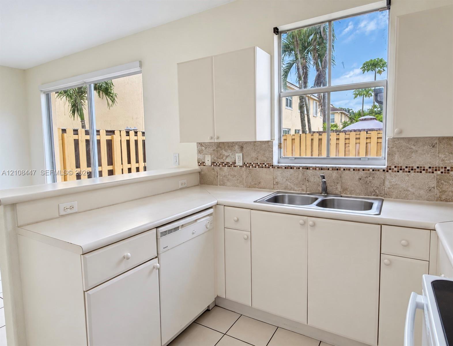 Kitchen featuring white cabinetry, solid-surface countertops, stone tile backsplash with decorative mosaic inlay, stainless steel dual-basin sink, and tile flooring