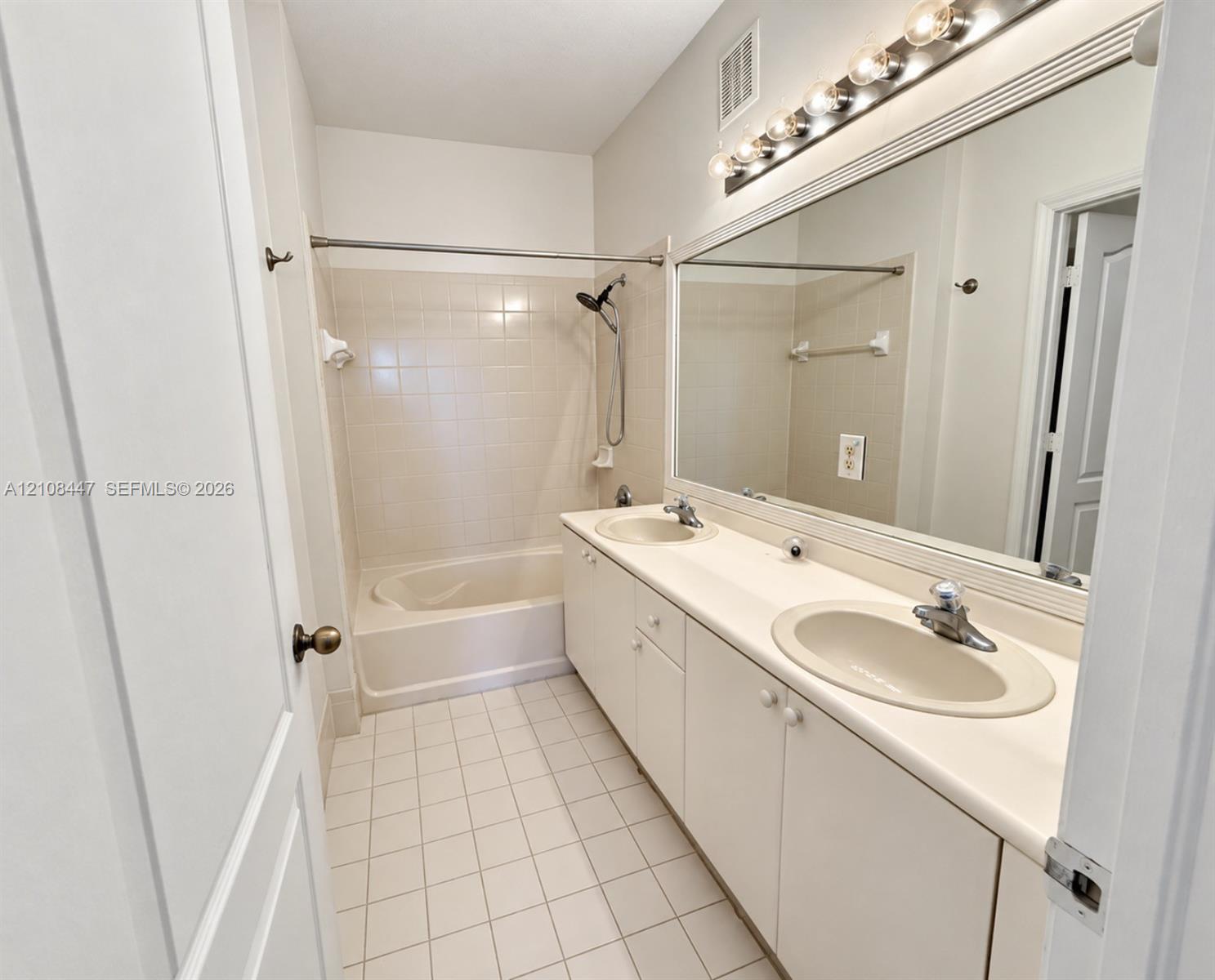 Bathroom featuring a dual-sink vanity with a solid-surface countertop, full-width vanity mirror, tile flooring, and a combined tub and shower enclosure with tile surround