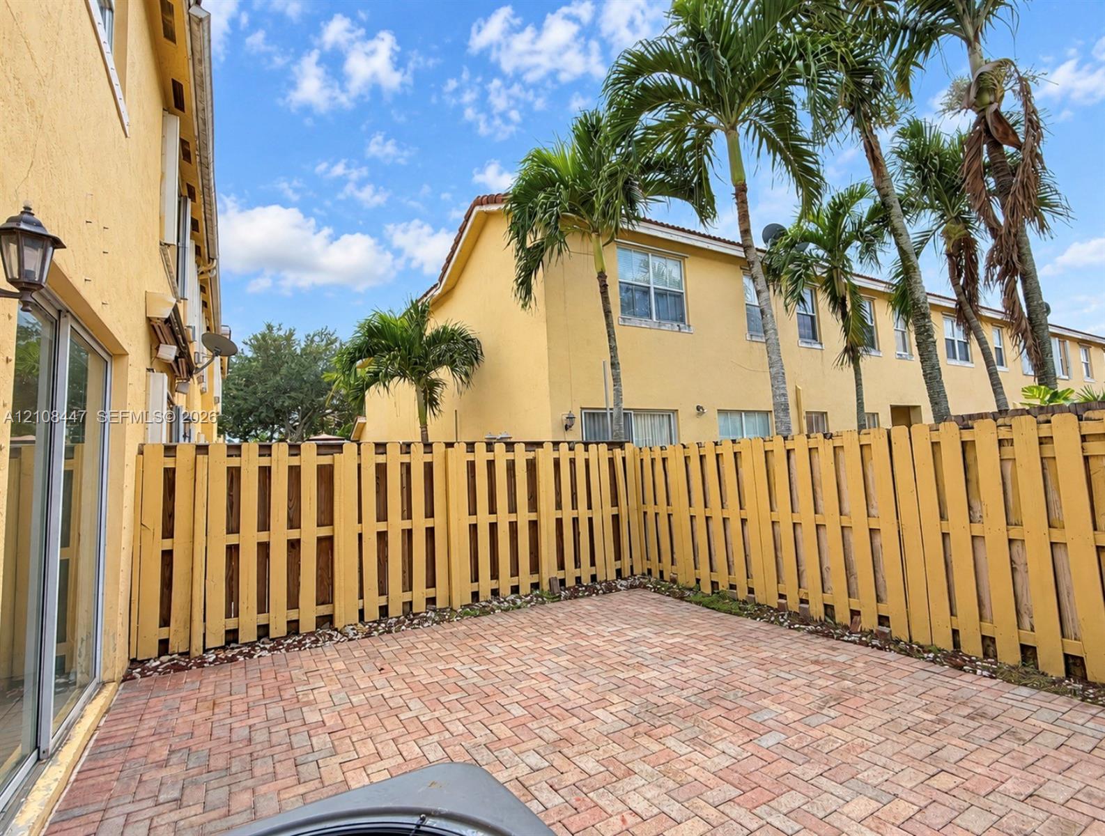 Private patio featuring herringbone-patterned brick pavers and a wooden privacy fence, complemented by mature palm trees