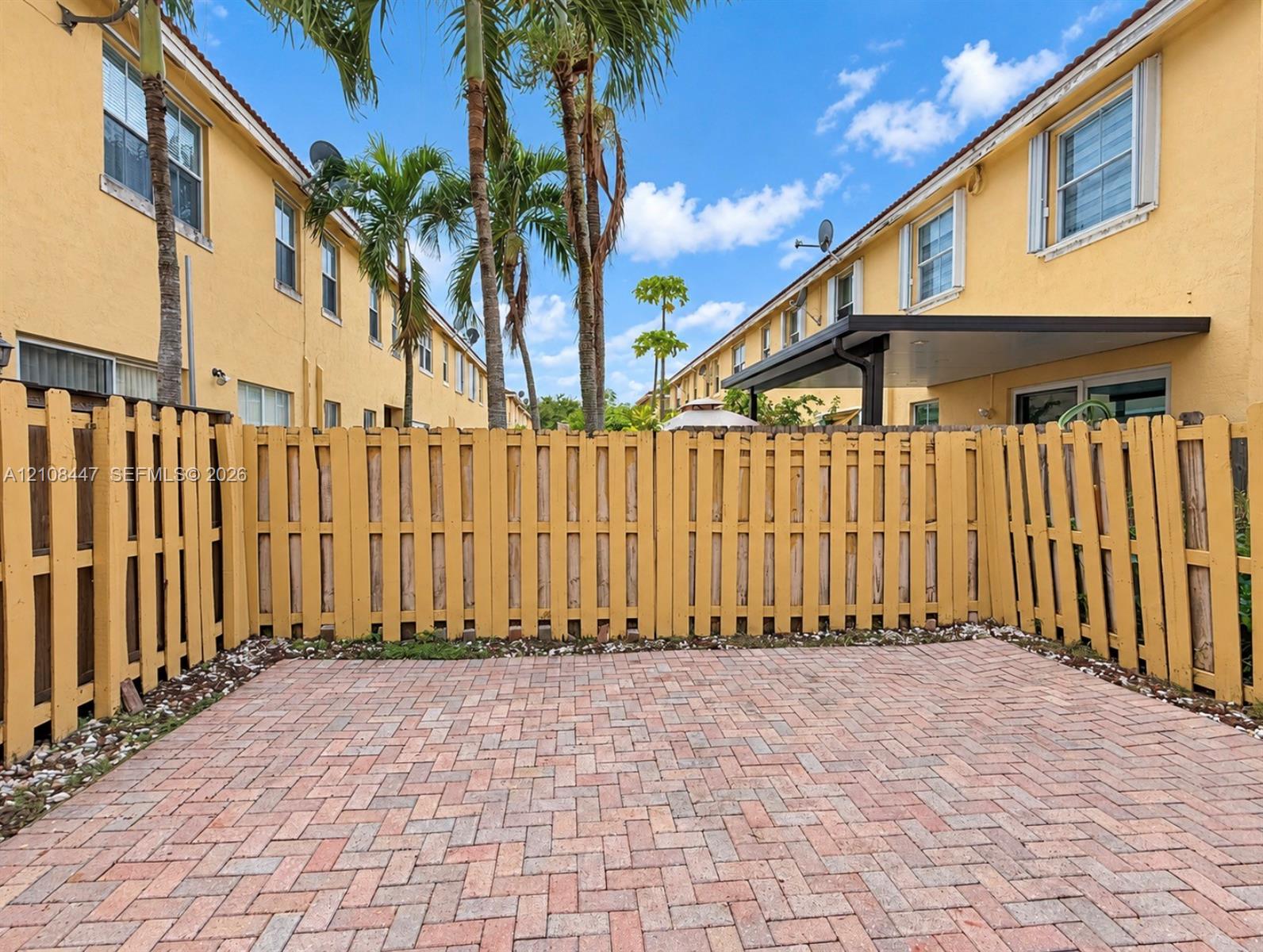 Private rear patio featuring herringbone-patterned pavers and a wood privacy fence, shaded by established palm trees