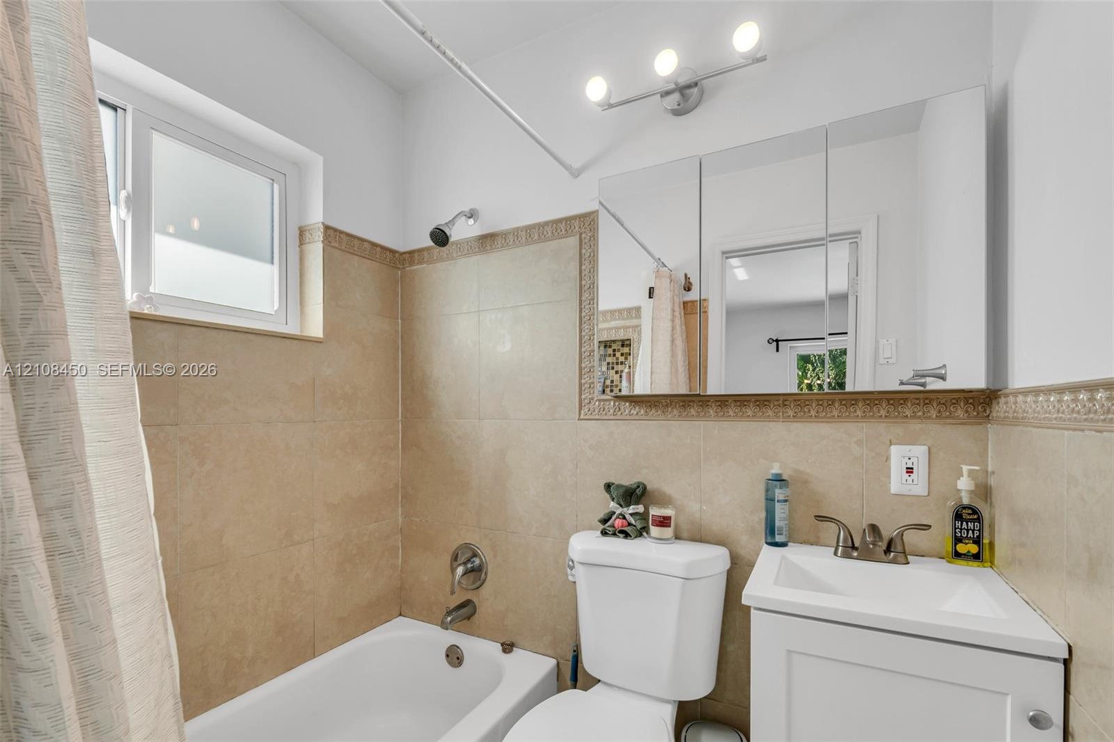 Bathroom featuring a white vanity cabinet, rectangular basin with dual-handle faucet, mirrored medicine cabinet, three-light fixture, and neutral tile surround with decorative border