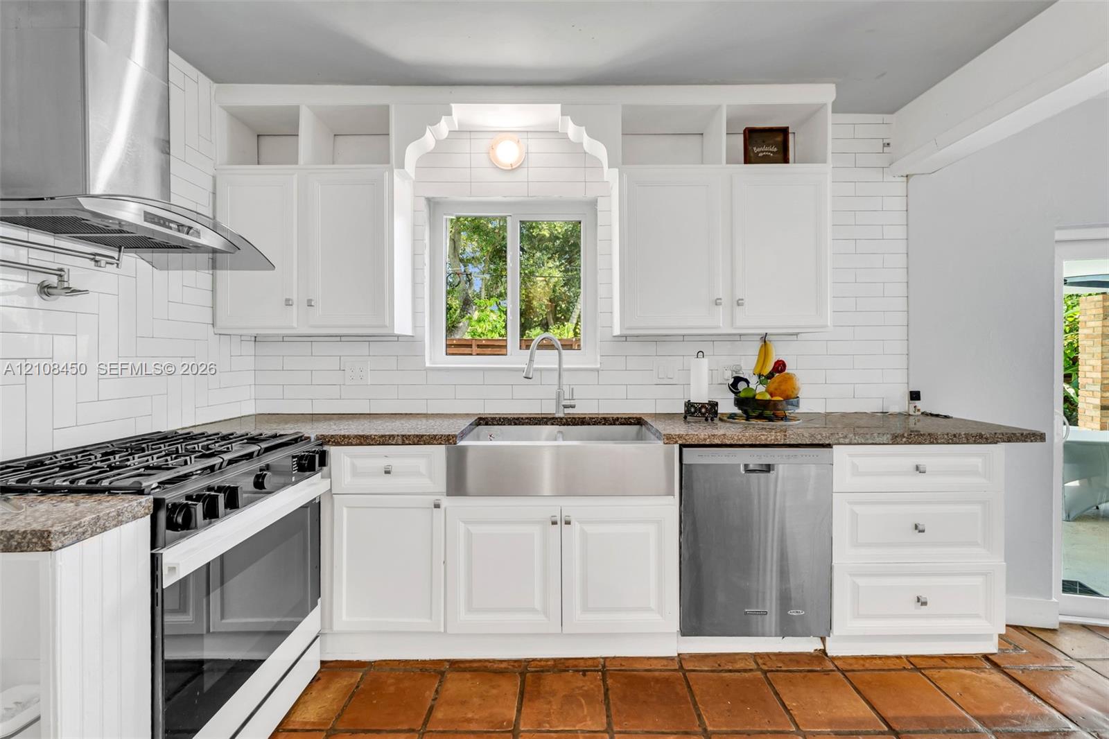 Kitchen featuring custom white cabinetry with decorative arch detailing, stainless steel farmhouse sink, herringbone-patterned subway tile backsplash, stone-surface countertops, and terracotta tile flooring