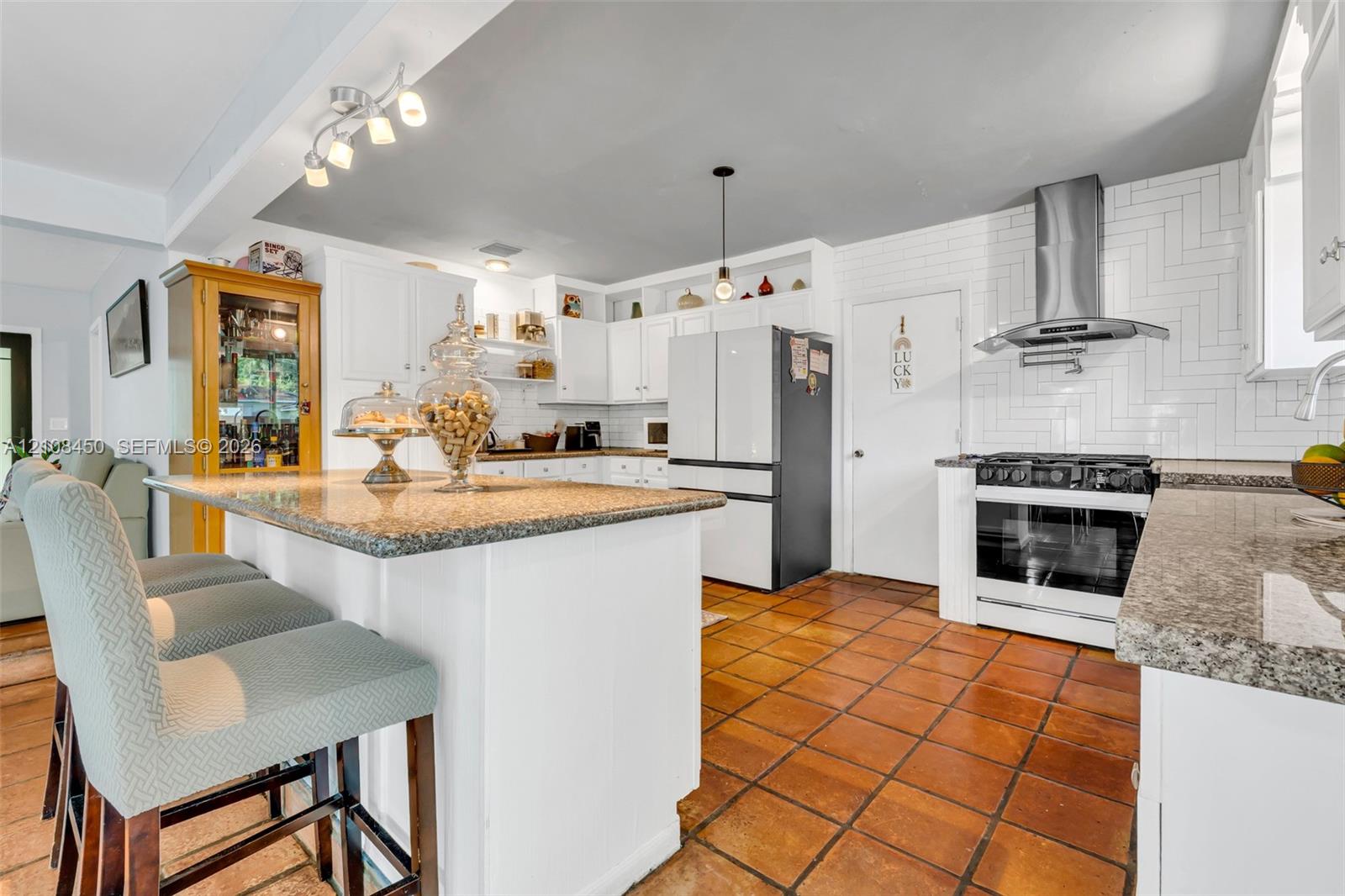 Kitchen featuring an island with a stone-finish countertop, white cabinetry, herringbone-patterned wall tile, stainless steel range hood, and terracotta-style flooring