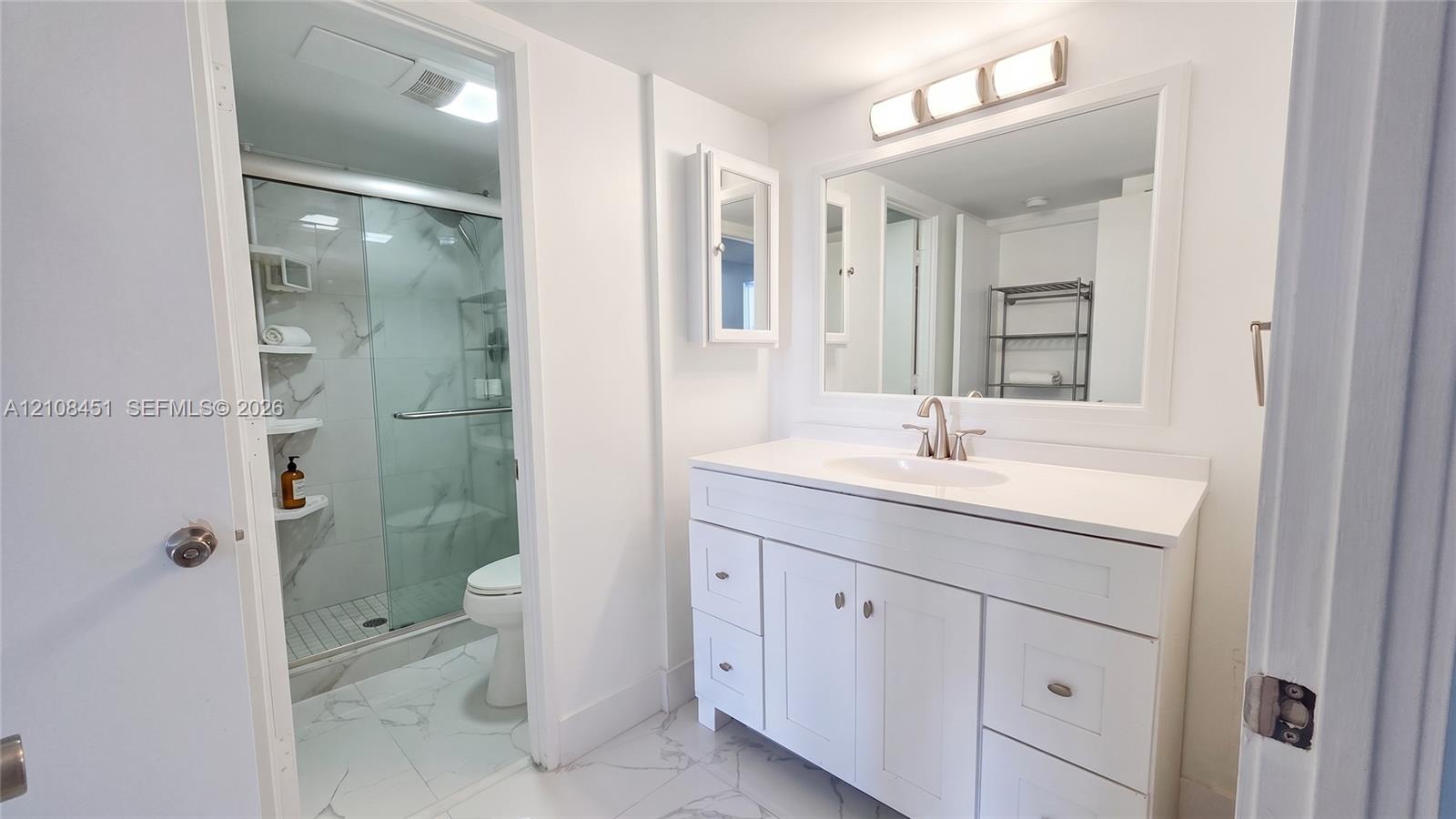 Bathroom featuring a white shaker-style vanity with stone-finish countertop, undermount sink, framed mirror with integrated lighting, and a glass-enclosed shower with tiled surround