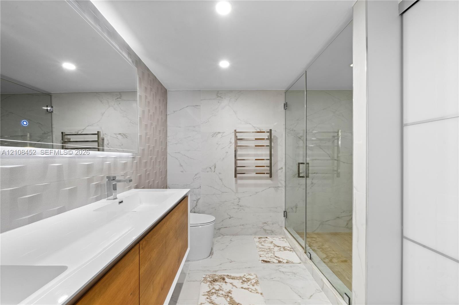 Floating wood-finish vanity with an integrated rectangular sink, wall-mounted metallic faucet, large frameless vanity mirror, textured three-dimensional wall tile, and glass-enclosed shower with floor-to-ceiling stone-look tile