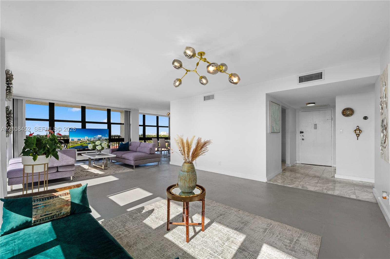 Open-concept living space featuring floor-to-ceiling black-frame windows, neutral tone flooring, and a multi-light brass-finish chandelier