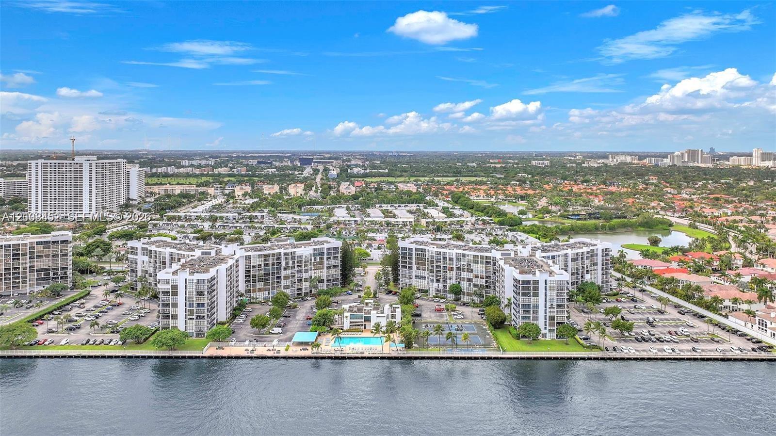 Aerial perspective of waterfront residential complex featuring expansive surface parking, on-site swimming pool, hard-surfaced tennis courts, and mature landscaping