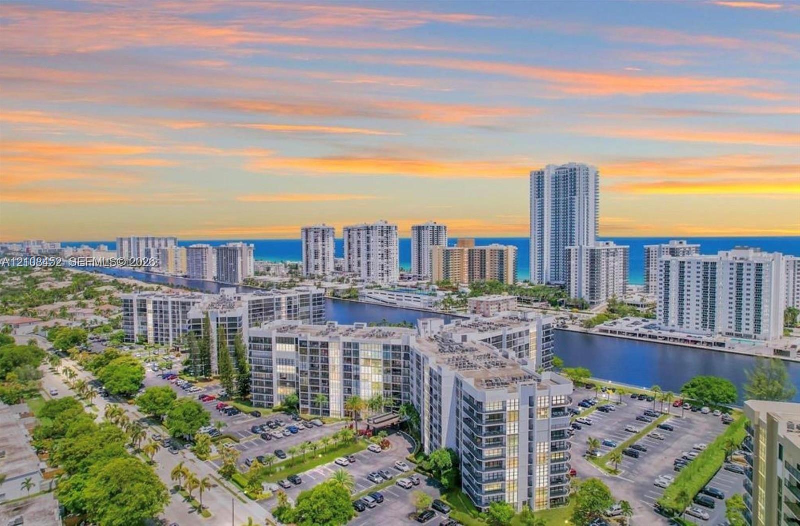 Aerial perspective featuring multi-story residential buildings, water canal, paved parking areas, and mature landscaping against a coastal horizon