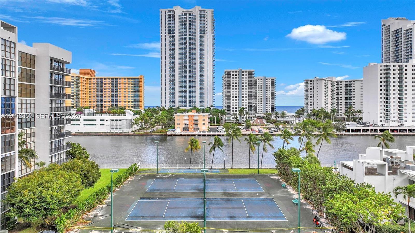 Outdoor tennis court, waterfront setting, paved perimeter walking path, manicured lawn areas, and mature palms