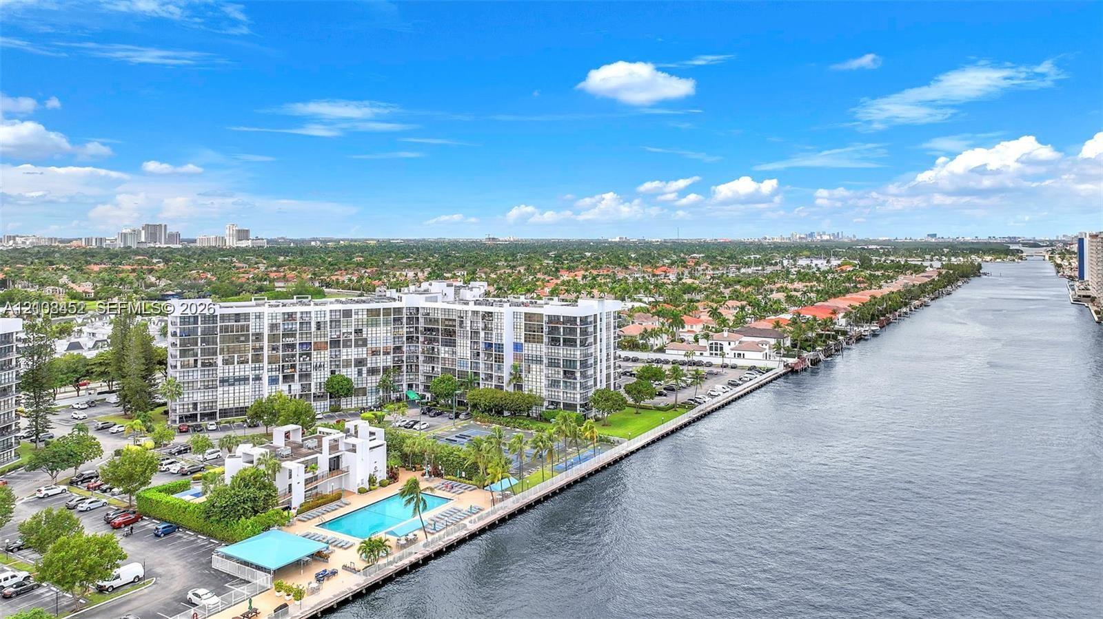 Waterfront mid-rise residential complex featuring a swimming pool, paved parking area, tennis court, and perimeter landscaping