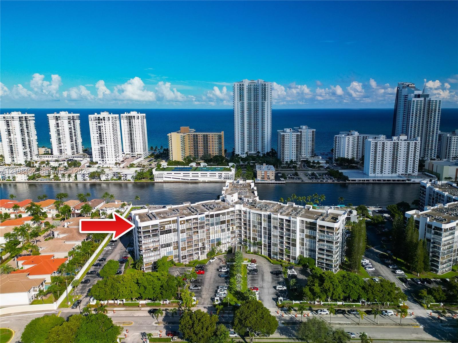Mid-rise multi-story residential building featuring exterior balconies, landscaped grounds, surface parking, and a waterfront location