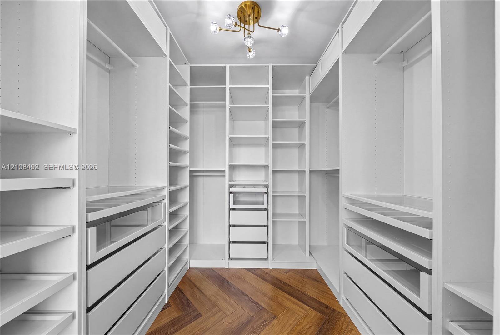 Walk-in closet featuring extensive built-in white cabinetry, multiple shelving tiers, integrated drawer units with glass-front detailing, chevron-patterned wood-finish flooring, and a multi-arm metallic light fixture