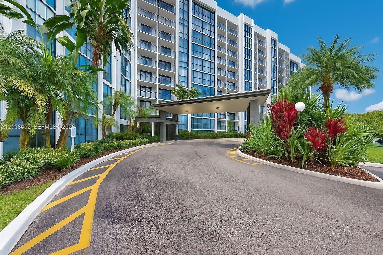 High-rise building exterior featuring a covered porte-cochre, glass facade, expansive balcony tiers, and landscaped grounds with mature palm trees