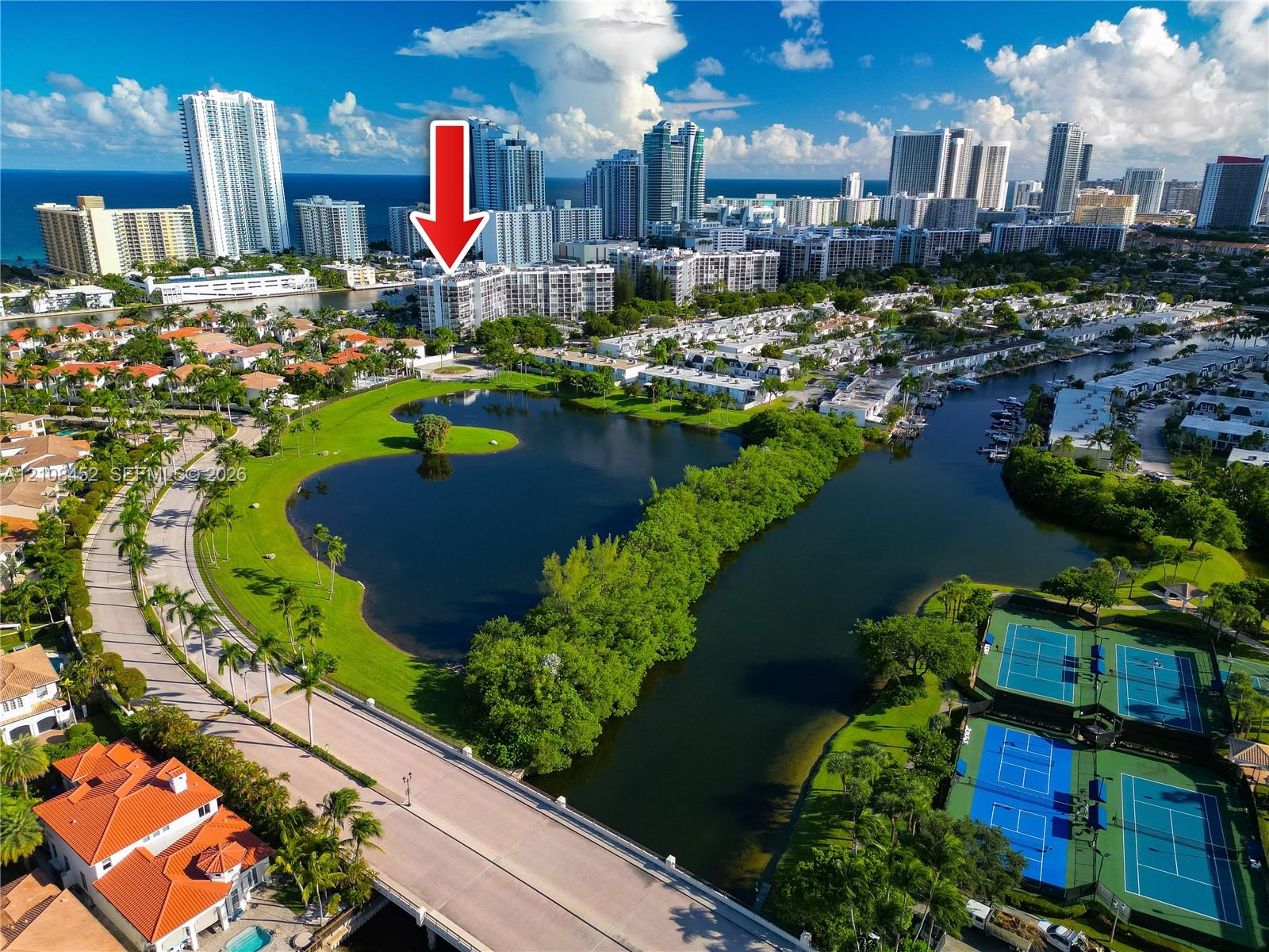 Aerial perspective of a multi-story residential building featuring proximity to a central lake, landscaped grounds, tennis courts, and coastal waterway