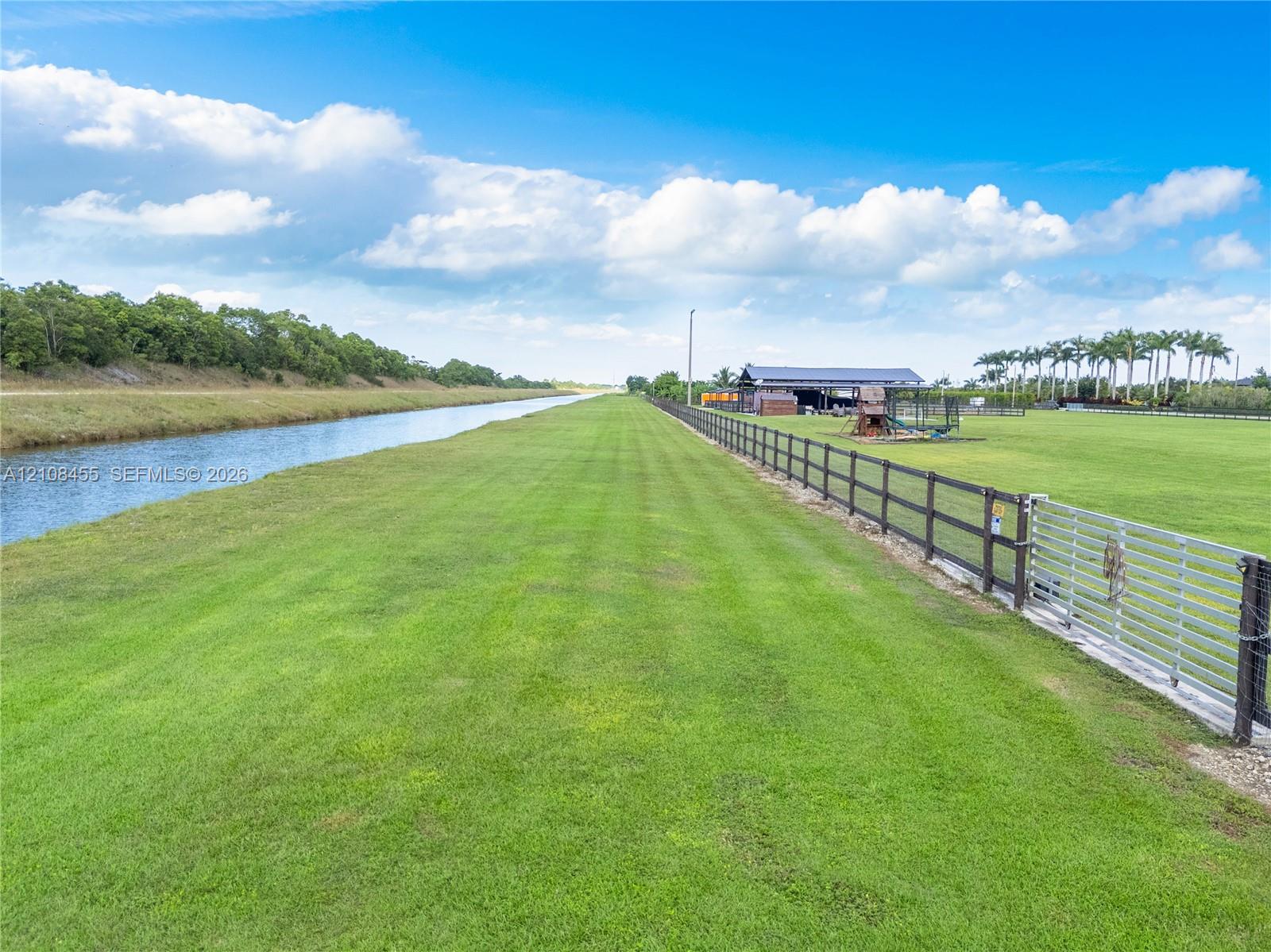Expansive acreage featuring a canal-front perimeter, manicured pastureland, a perimeter post-and-rail fence, a metal gate, and an open-sided pole barn structure