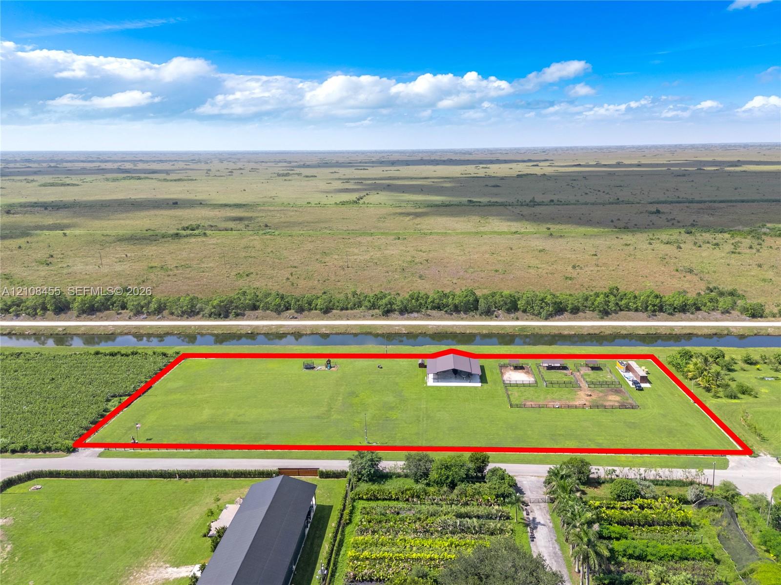 Expansive acreage featuring a metal-roof structure, fenced outdoor enclosures, a canal-front perimeter, and open grassland