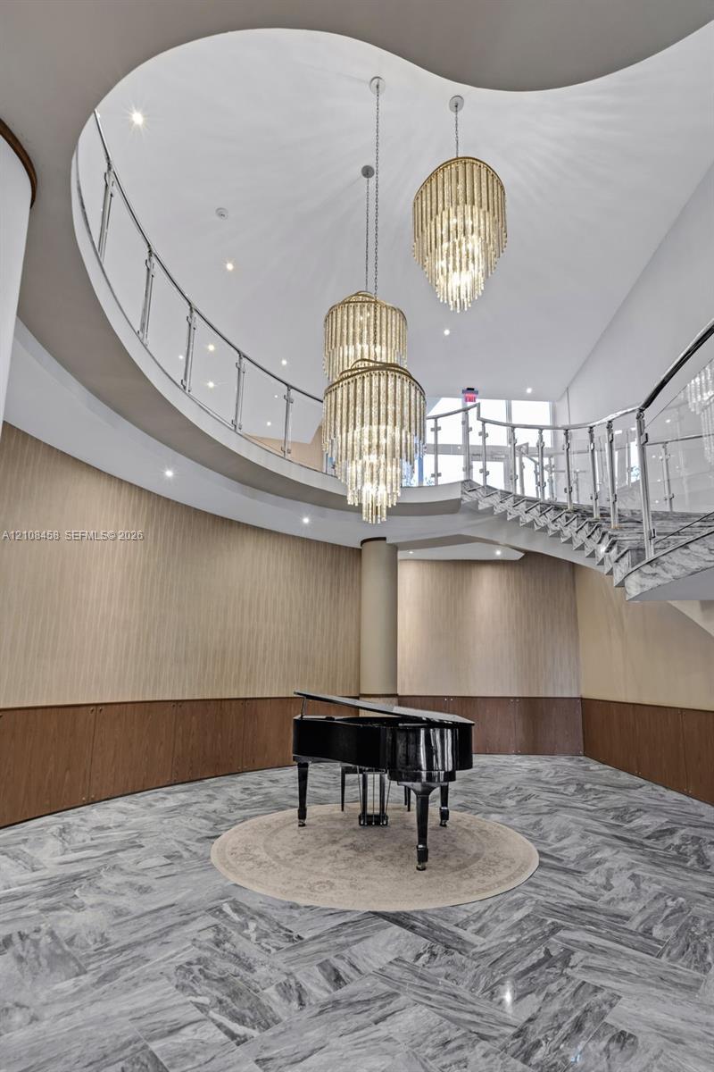 Grand foyer featuring a sweeping curved staircase with polished metal railings, tiered crystal chandeliers, stone flooring in a herringbone pattern, and wood-paneled walls