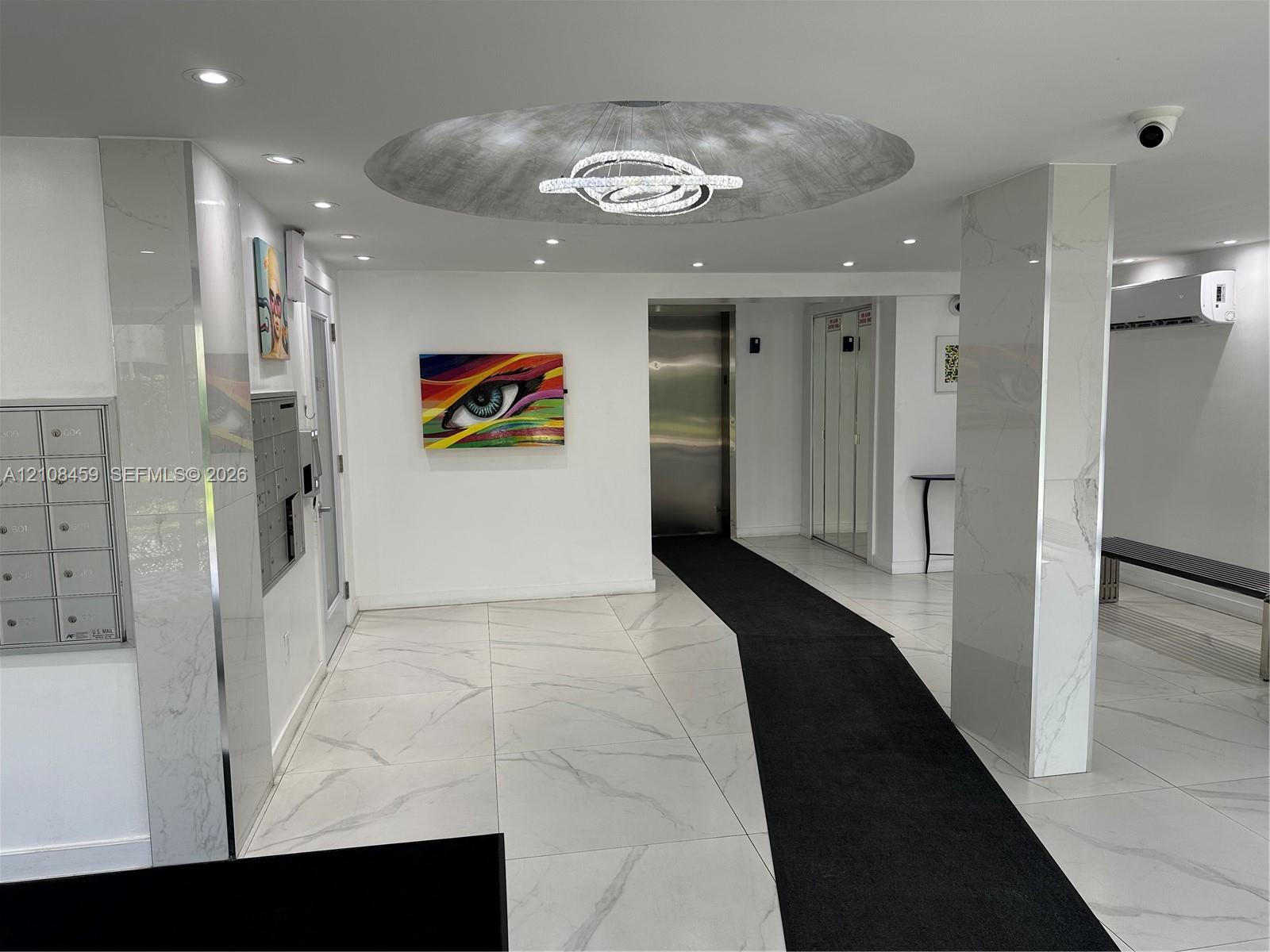 Lobby interior featuring large-format stone-finish floor tiling, polished stone-clad columns, recessed lighting, a modern sculptural ceiling fixture, and stainless steel elevator doors