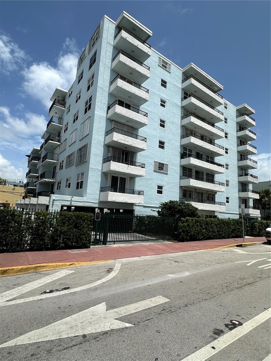 Multi-story residential building featuring a light blue stucco exterior, stacked private balconies with black metal railings, and a gated ground-level parking entrance Multi-story residential building featuring a light blue stucco exterior, stacked private balconies with black metal railings, and a gated ground-level parking entrance
