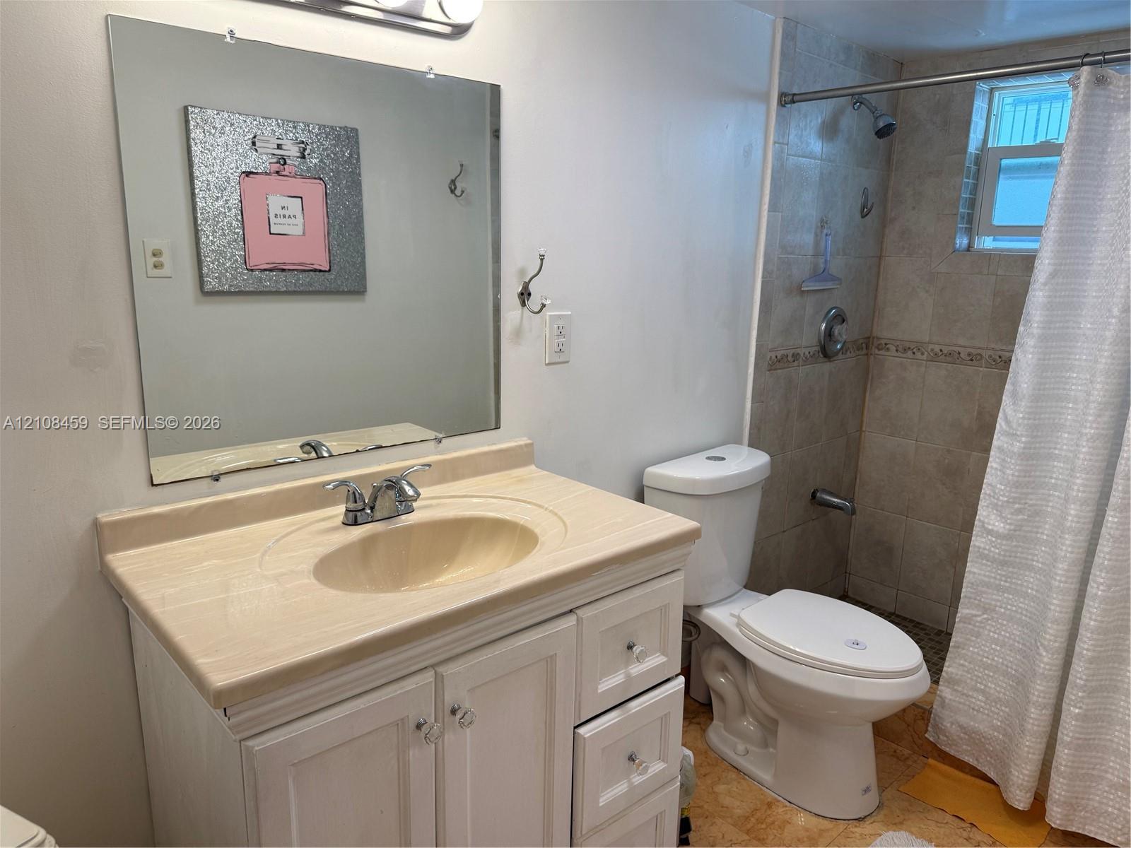Vanity with stone-finish countertop, integrated sink, and white cabinetry, paired with a square wall-mounted mirror, ceramic tile shower surround, and chrome shower fixtures Vanity with stone-finish countertop, integrated sink, and white cabinetry, paired with a square wall-mounted mirror, ceramic tile shower surround, and chrome shower fixtures